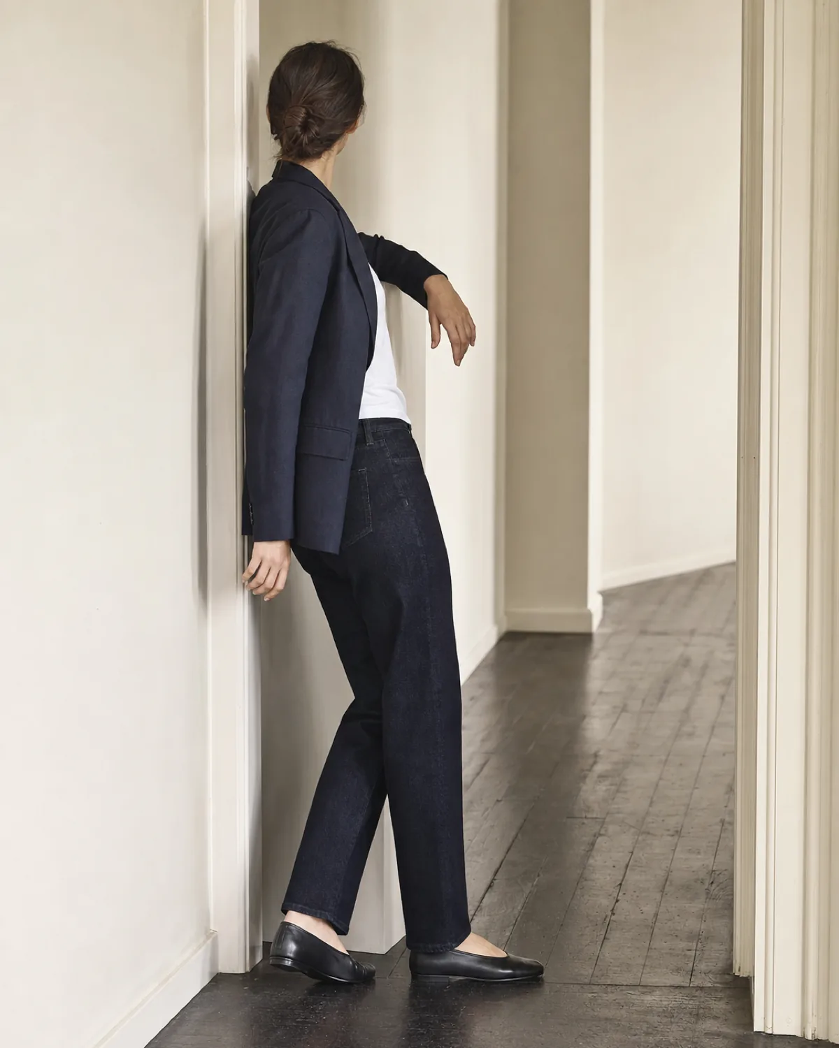 A woman seen from behind leaning one shoulder in a plain doorway, wearing dark indigo straight-leg jeans, a navy blazer, a white tee and black loafers