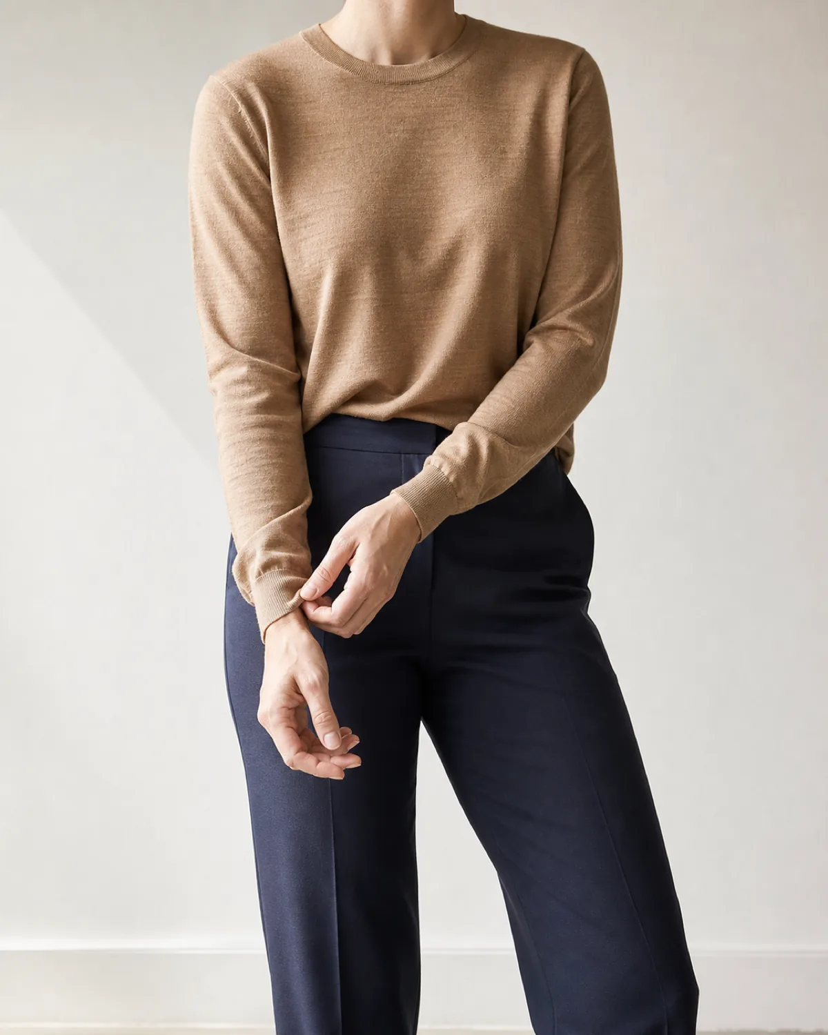 A camel fine gauge knit worn untucked over navy tailored trousers, cropped from chin to knee, hands adjusting the cuff